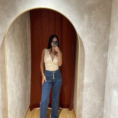 Mulher a vestir top creme de decote em V e calças de ganga azuis, em frente a um espelho oval com moldura dourada