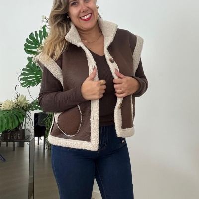 Colete castanho com forro branco e botões, usado por mulher em ambiente interior, com plantas ao fundo.