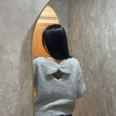 Mulher de costas a usar camisola cinzenta felpuda com laço grande e recorte em forma de coração nas costas, em frente a parede texturizada cinzenta com arco