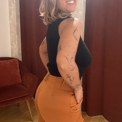 Mulher de blusa preta e calças castanhas em sala com cortinas e poltrona castanhas