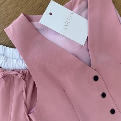 Colete rosa com botões pretos e etiqueta CAMELIA, com calça rosa ao lado sobre madeira