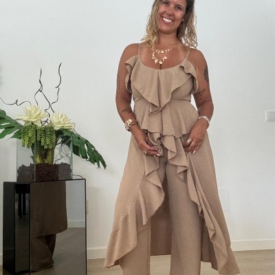 Mulher a usar vestido bege com folhos e calças largas, ao lado de planta decorativa num móvel espelhado.