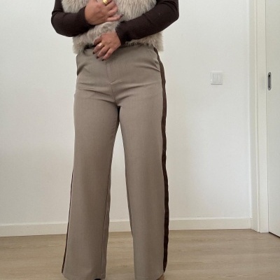 Pessoa vestindo calças bege com faixa lateral castanha, blusa de malha castanha e colete felpudo bege, em sala com chão de madeira e parede branca.