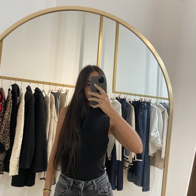 Mulher em top preto e jeans à frente de espelho dourado com roupas penduradas ao fundo
