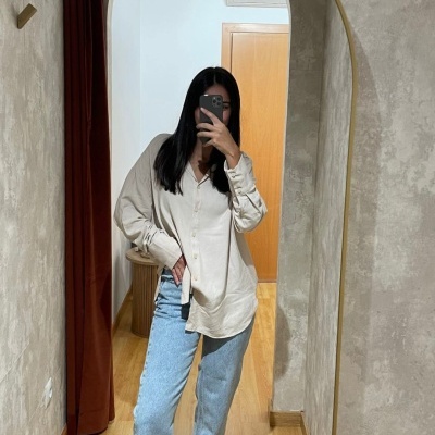 Pessoa com camisa bege, calças jeans azuis claras e ténis brancos em frente a espelho oval com moldura dourada.