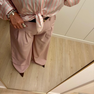 Pessoa a usar camisa de riscas rosa e branco com nó, calças largas rosa claro e acessórios prateados diante de um espelho