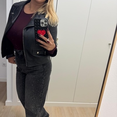 Mulher com casaco preto e calças pretas com padrão brilhante tira selfie com telemóvel em espelho