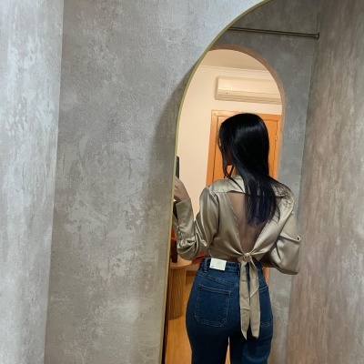 Mulher com blusa bege acetinada e calças de ganga azuis vista de costas num espelho