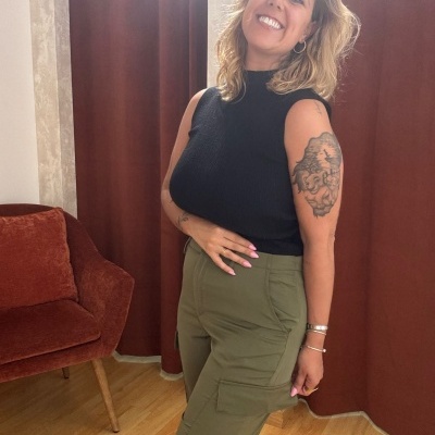 Mulher com calças verdes cargo e camisola preta, com cadeira e cortina castanhas ao fundo