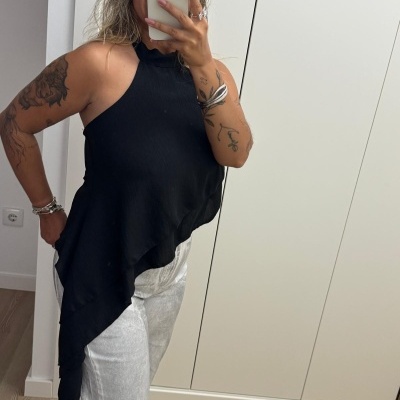 Top feminino preto assimétrico com calças claras e pulseiras, retrato selfie em espelho interior.