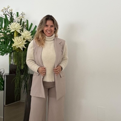 Mulher com colete bege e camisola de gola alta creme ao lado de vaso com plantas e flores