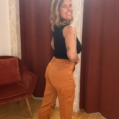 Pessoa com calças de cargo castanhas e top preto num ambiente interior com cortinas e cadeira castanhas