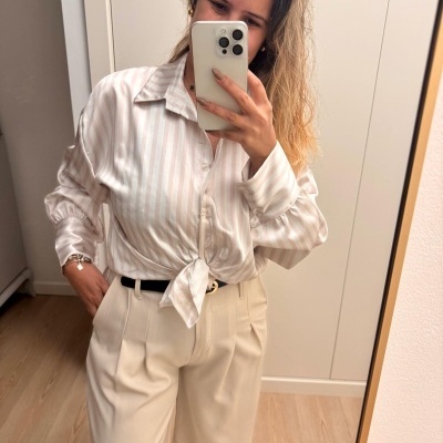 Mulher com camisa riscada clara amarrada na cintura e calças bege largas junto a um espelho