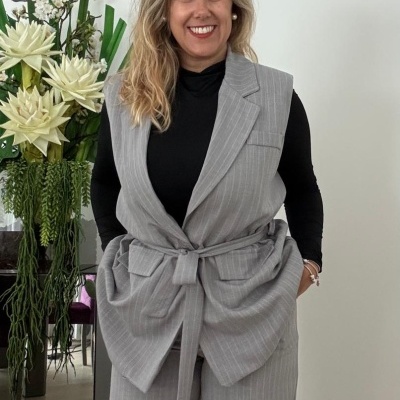 Conjunto cinzento de riscas com colete e calças, blusa preta, mulher num ambiente interior com plantas decorativas.