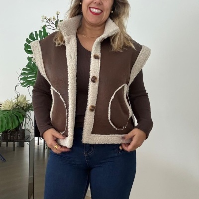 Colete castanho com pelo creme e botões, sobre camisola castanha e jeans azul, com plantas ao fundo.