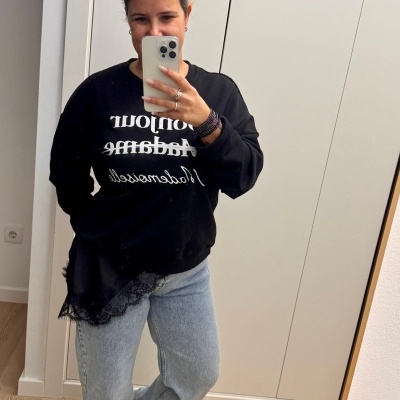 Mulher a tirar selfie de corpo inteiro com sweat preto, calças de ganga azul e sapatos branco e preto em quarto com armário branco.
