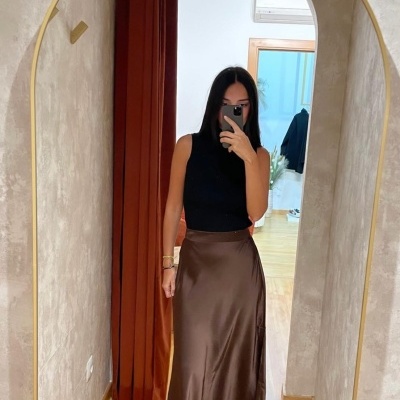 Mulher com top preto e saia castanha acetinada frente a espelho dourado.