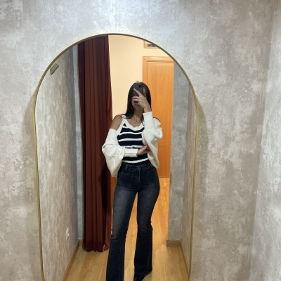 Espelho oval com moldura dourada refletindo mulher com roupa preta e branca num corredor