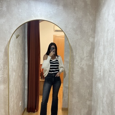 Espelho oval com moldura dourada numa parede cinza e pessoa vestindo blazer branco e jeans pretos na reflexão