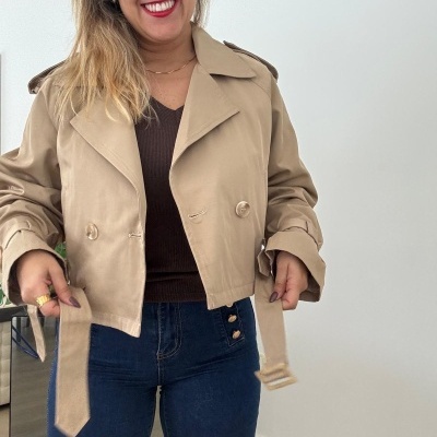 Mulher a vestir casaco curto bege com cinto, camisola castanha e calças de ganga azul.