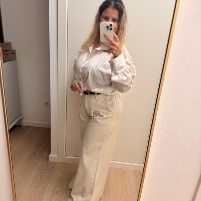 Pessoa vestindo camisa branca com riscas e calças largas bege, tirando selfie em espelho