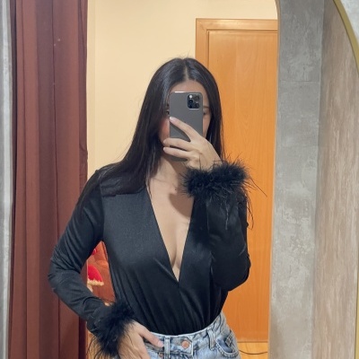 Pessoa com blusa preta de decote e penas nas mangas, calças de ganga azuis claras, selfie em espelho em quarto interior