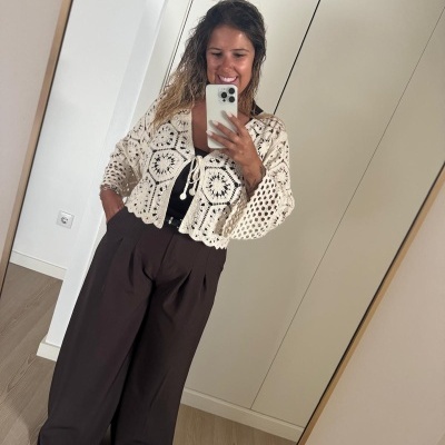 Mulher com casaco de croché creme e calças largas castanhas a tirar selfie num quarto