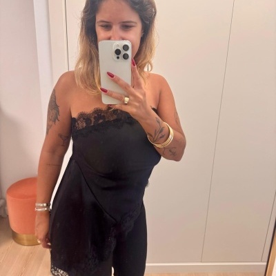 Mulher veste top preto com renda, calças pretas e acessórios, tirando selfie no espelho.