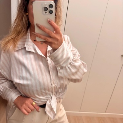 Pessoa a usar camisa de riscas brancas e bege com nó na cintura e calças bege, tirando foto de espelho em divisão com armários brancos.