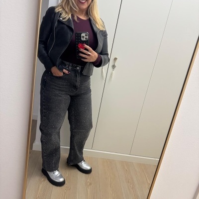 Mulher vestindo casaco preto e calças de ganga cinza com pontos, sapatos prateados, tirando selfie em espelho de quarto