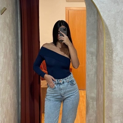 Mulher com blusa azul escuro ombro único e calças de ganga claras em espelho com moldura dourada.