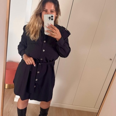 Mulher a usar vestido preto com botões e botas pretas em frente a um armário branco