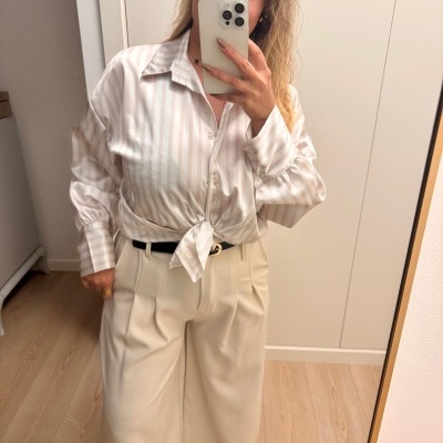 Mulher a tirar selfie com camisa acetinada riscada e calças largas bege numa divisão minimalista.