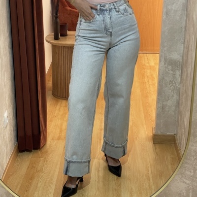 Pessoa a usar calças de ganga azul clara dobradas com top rosa e sapatos pretos, refletida num espelho de corpo inteiro num quarto com chão de madeira.