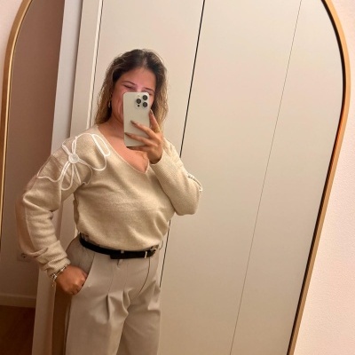 Mulher a tirar selfie em espelho redondo dourado, vestindo camisola bege com padrão branco e calças bege