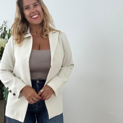Mulher com blazer branco, top castanho claro e calças de ganga azul em interior com parede branca e plantas