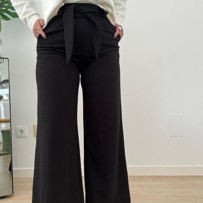 Calças pretas de tecido fluido com corte largo e faixa na cintura, combinadas com camisola branca, em ambiente interior