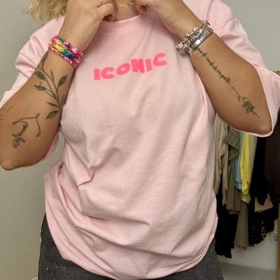 T-shirt rosa com a palavra ICONIC em letras fluorescentes, usada por pessoa com pulseiras e tatuagens, em ambiente interior com roupas ao fundo.