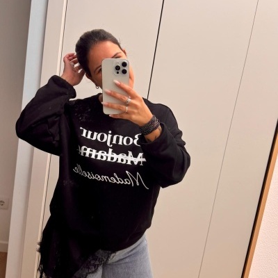 Pessoa a usar sweatshirt preta com estampado branco, segurando telemóvel prata à frente do espelho em ambiente interior.