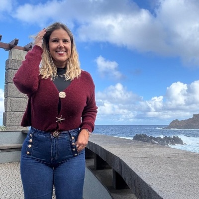 Mulher a vestir cardigan bordô e calças de ganga perto do mar com céu azul e nuvens.