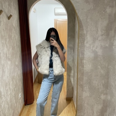 Mulher de pé vestindo colete de pelo branco e calças de ganga, refletida em espelho oval