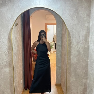 Pessoa a usar vestido comprido preto com sandálias pretas, refletida em espelho oval com moldura dourada numa divisão com parede cinzenta e chão de madeira