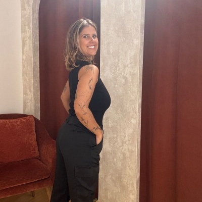 Mulher de roupa preta com tatuagens em ambiente interior com cortinas castanhas e sofá vermelho