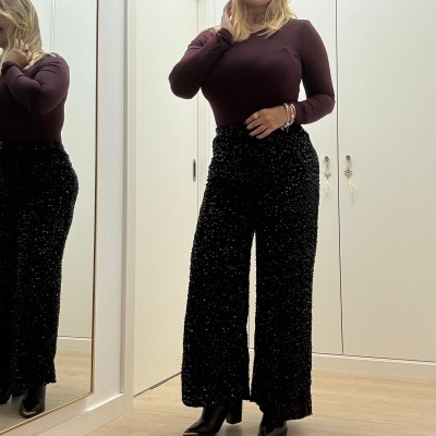 Mulher com camisola castanha e calças pretas texturizadas em interior com espelho e chão de madeira.