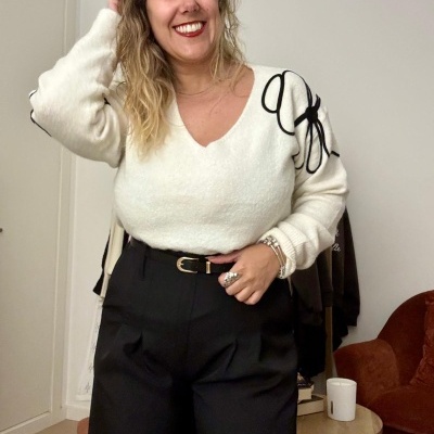 Mulher a usar camisola branca de malha com padrão preto nos ombros e calças pretas com cinto, em interior com sofá castanho.