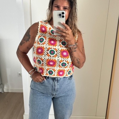 Top crochet colorido com quadrados e calças jeans azuis em selfie dentro de um quarto.