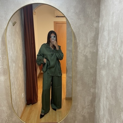 Mulher a vestir conjunto verde acetinado e a tirar foto num espelho oval numa sala com paredes cinzentas e chão de madeira