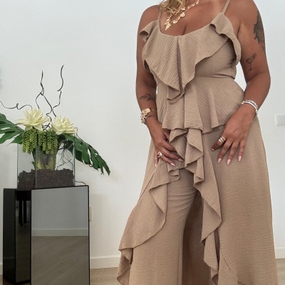 Mulher vestindo conjunto bege com textura e folhos, pulseiras e colar dourados, em ambiente interior com planta e móvel refletor.