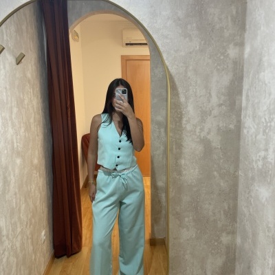 Mulher a usar calças largas e colete azul claro em frente a um espelho oval com moldura dourada