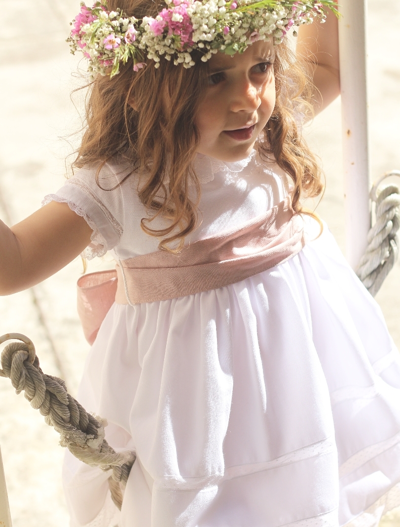Criança com vestido branco e faixa rosa numa baloiço com coroa de flores.
